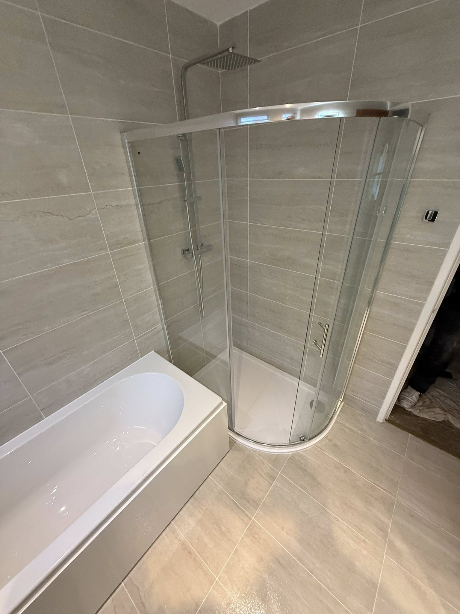 Newly installed modern shower enclosure next to a white bathtub with beige wall and floor tiling.