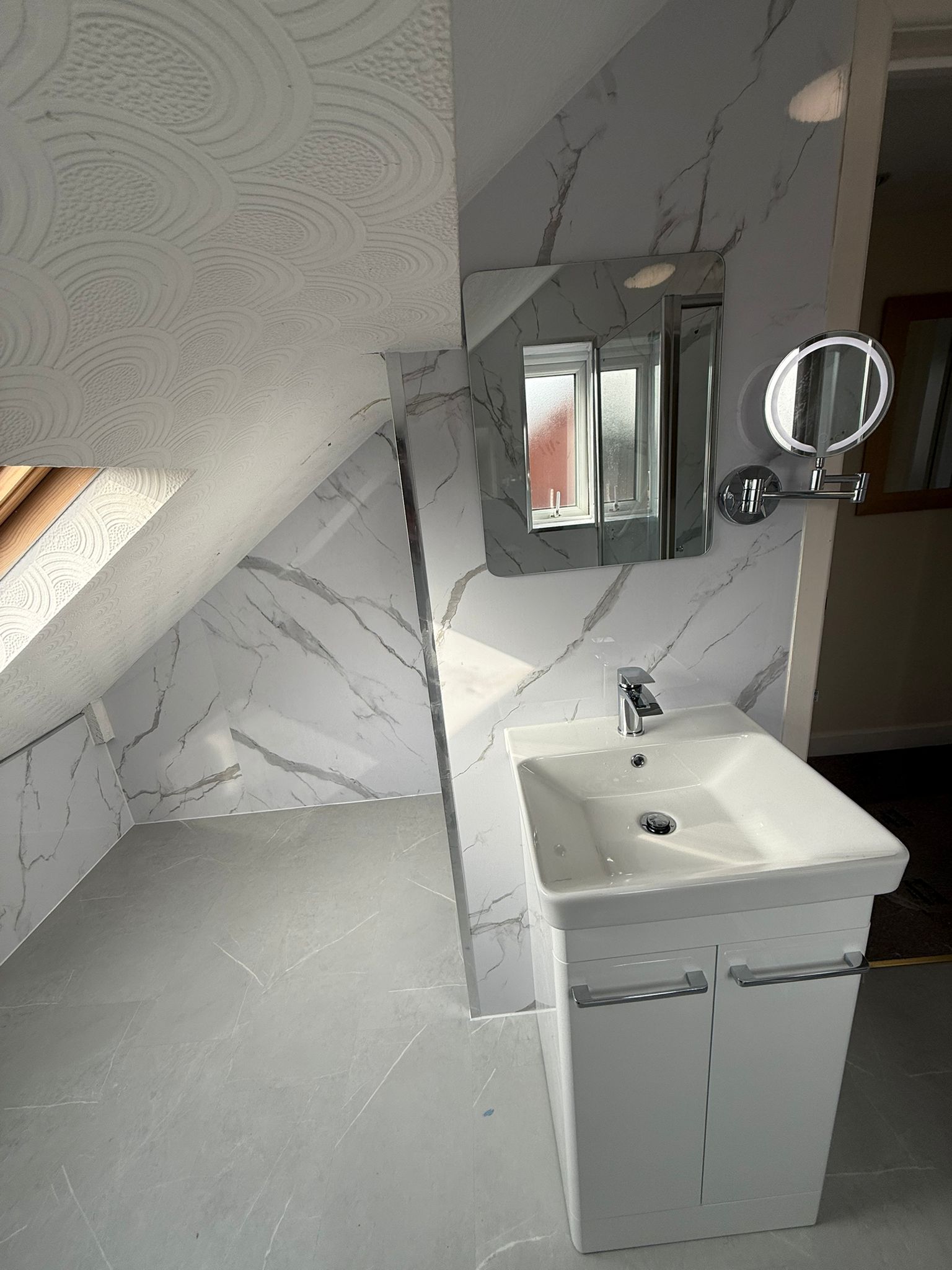 Modern bathroom vanity with white sink, chrome faucet, and illuminated mirror against white marble-effect wall tiles.